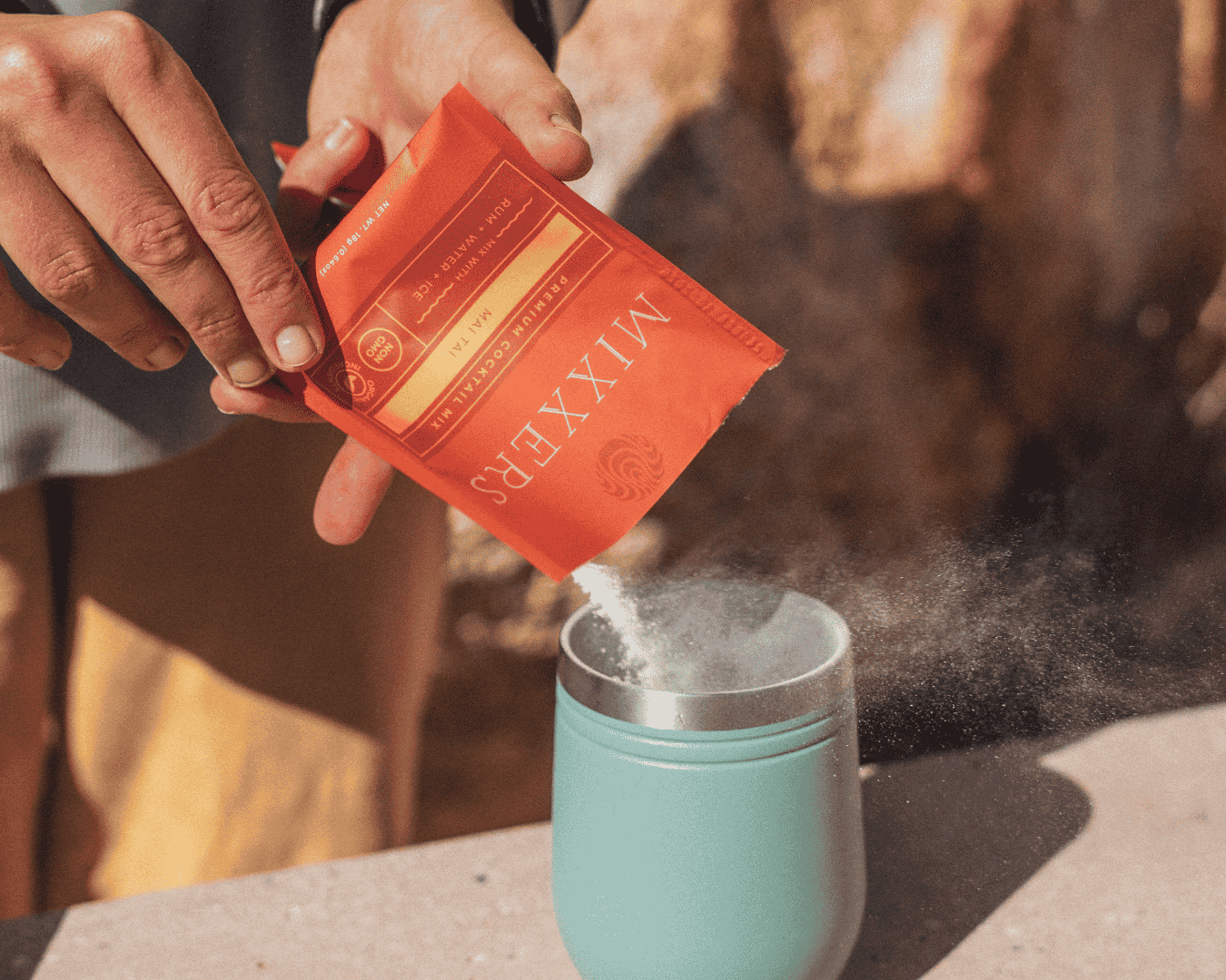 Person pouring a red packet of Mixxers into a teal mug with steam rising.